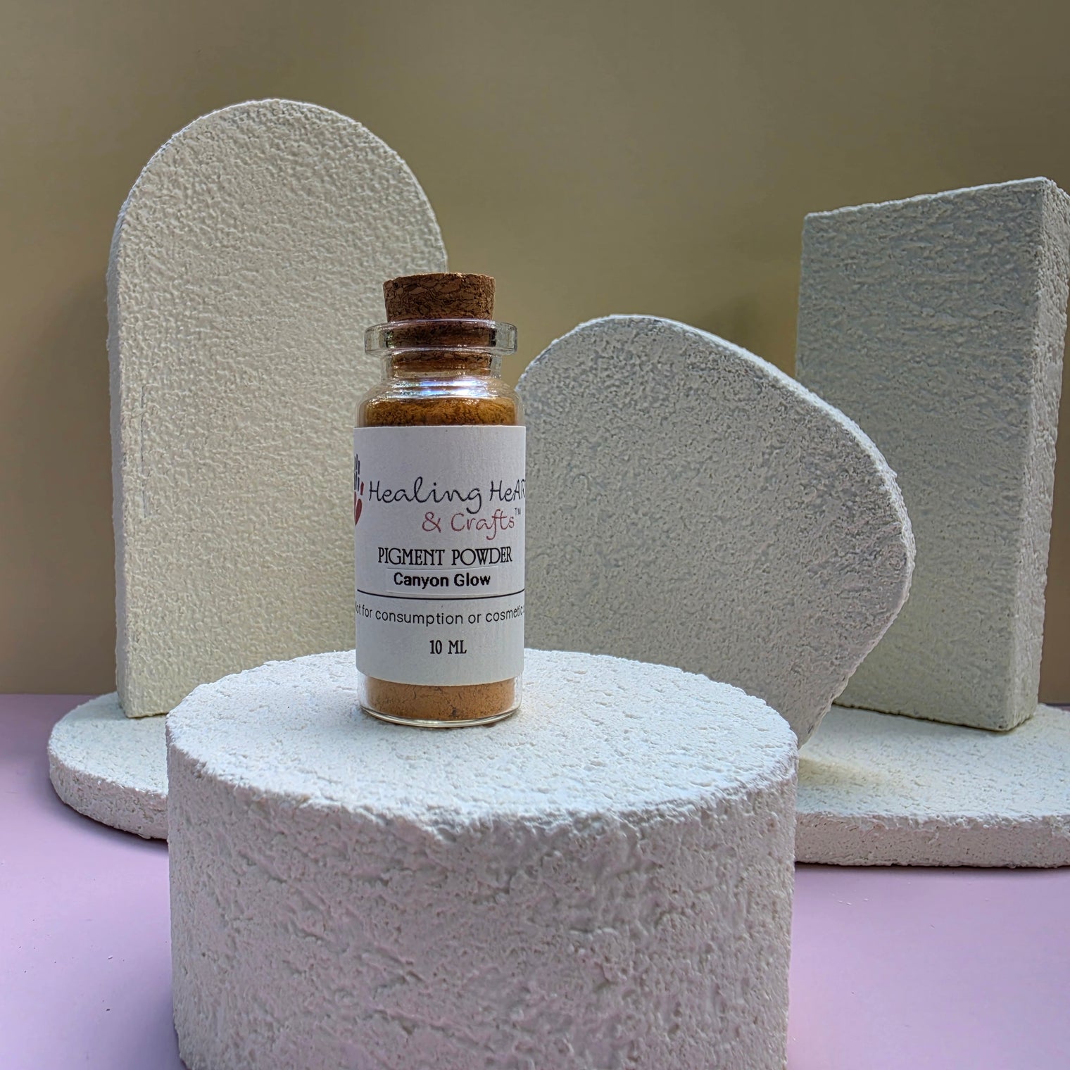 A small glass bottle of Healing Hearts & Crafts Canyon Glow Paint pigment powder with a cork stopper, displayed on a textured white cylindrical pedestal. The background features abstract, textured white geometric shapes against a beige wall, with a light purple surface at the bottom.