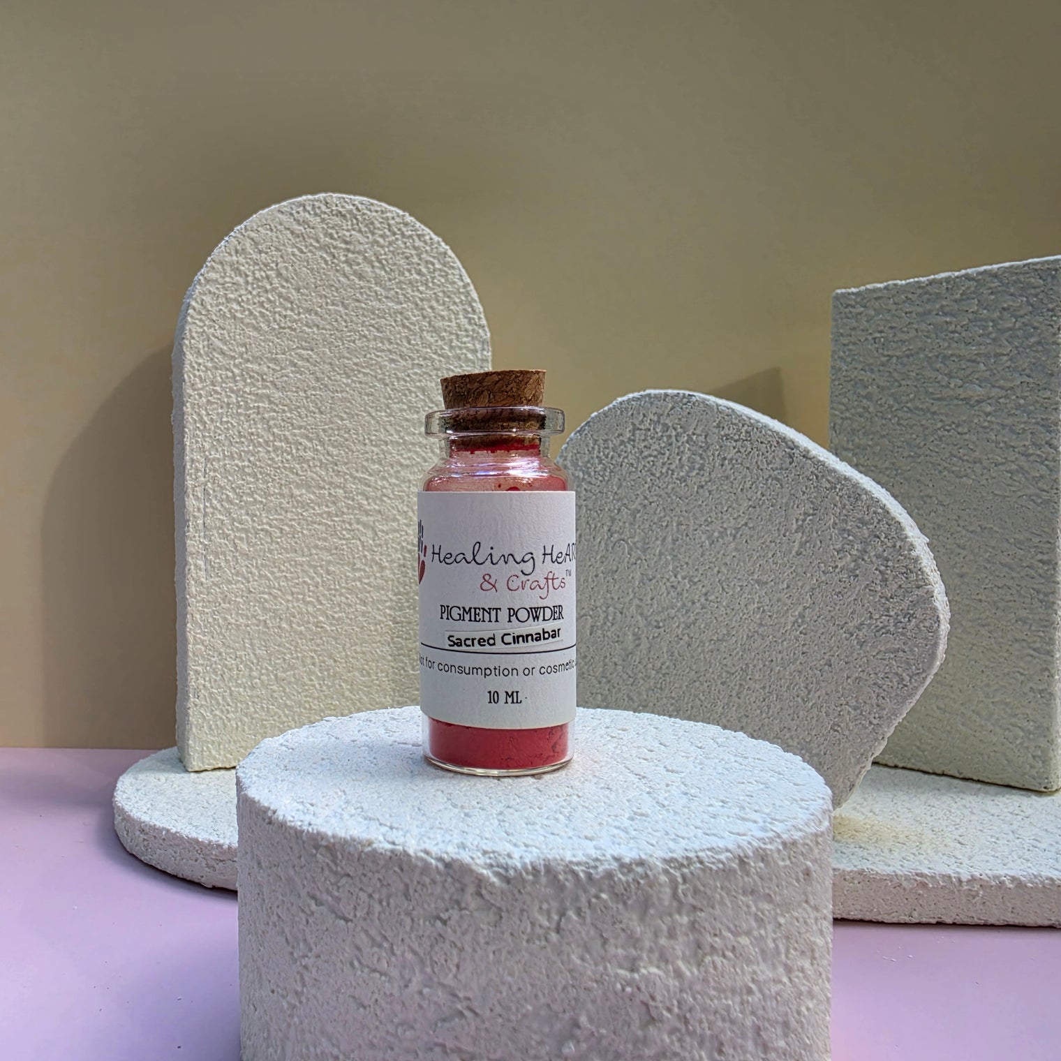 A small glass vial of Healing Hearts & Crafts pigment powder sits on a textured white cylindrical pedestal. The vial contains a vibrant pink pigment powder called Sacred Cinnabar and is sealed with a brown cork stopper featuring a white label.