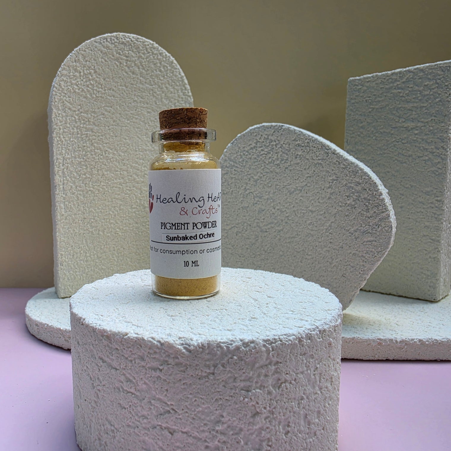A small glass vial of Healing Hearts & Crafts Paint Pigment Powder in sunbaked Ochre, a vial containing golden-brown pigment powder, displayed on a textured white foam pedestal against a beige background.