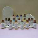A collection of Healing Hearts & Crafts paint pigment powder samplers. Small glass bottles filled with colorful powder arranged in front of two textured white display stands on a pale pink surface against a beige wall. The bottles have cork stoppers and white labels reading 'Pigment Powder' and various other color names.
