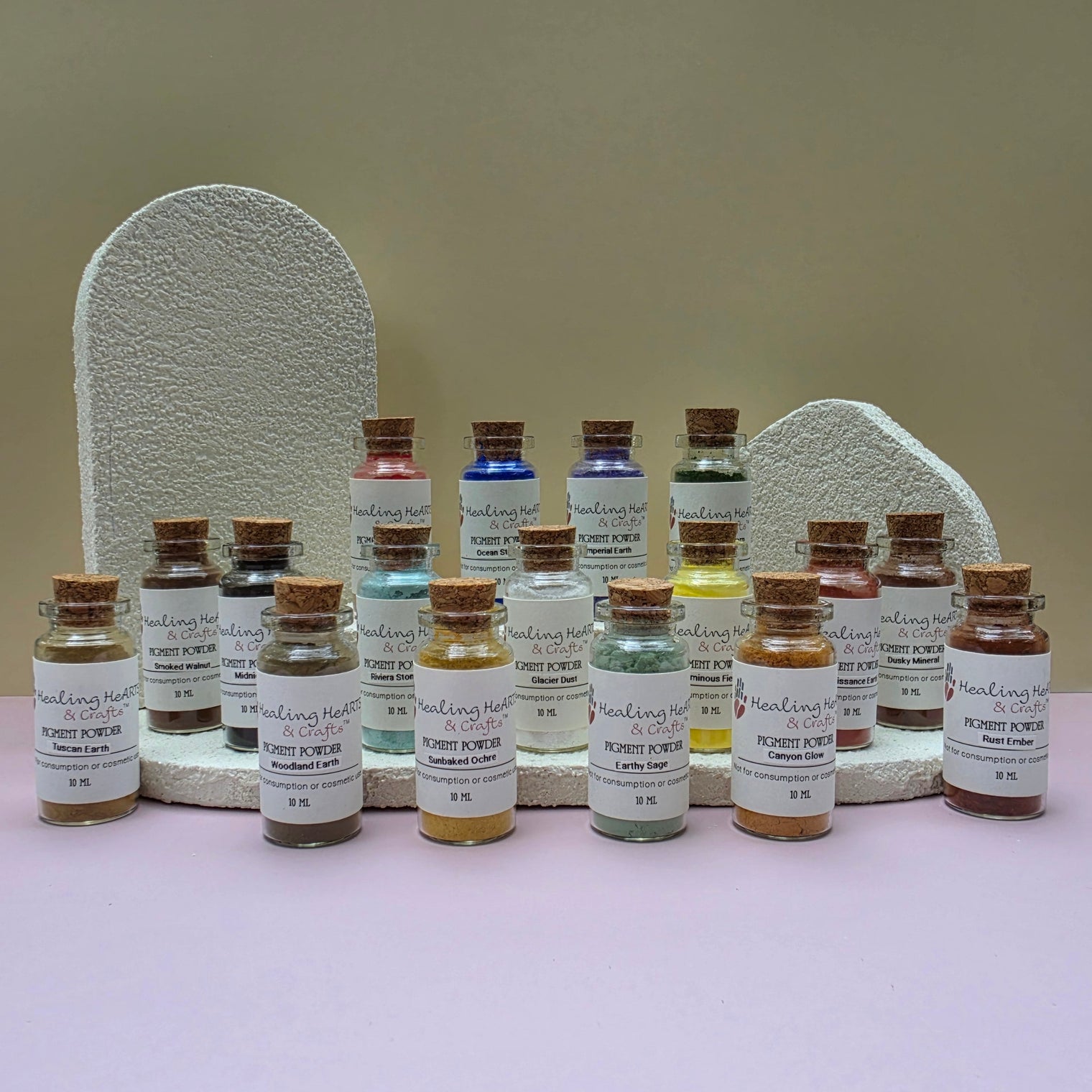 A collection of Healing Hearts & Crafts paint pigment powder samplers. Small glass bottles filled with colorful powder arranged in front of two textured white display stands on a pale pink surface against a beige wall. The bottles have cork stoppers and white labels reading 'Pigment Powder' and various other color names.