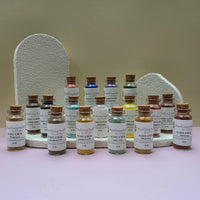A collection of Healing Hearts & Crafts paint pigment powder samplers. Small glass bottles filled with colorful powder arranged in front of two textured white display stands on a pale pink surface against a beige wall. The bottles have cork stoppers and white labels reading 'Pigment Powder' and various other color names.