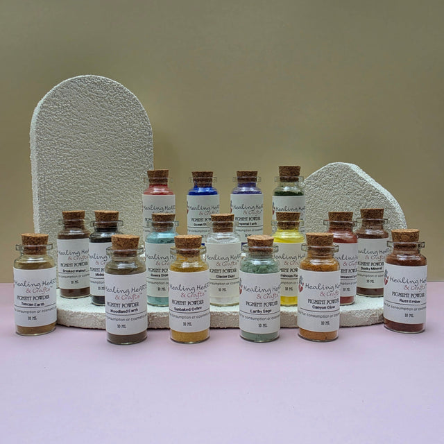 A collection of Healing Hearts & Crafts paint pigment powder samplers. Small glass bottles filled with colorful powder arranged in front of two textured white display stands on a pale pink surface against a beige wall. The bottles have cork stoppers and white labels reading 'Pigment Powder' and various other color names.