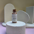 A small glass bottle of Healing Hearts & Crafts Paint Pigment Powder in Dusky Mineral displayed on a textured white foam pedestal, with matching white foam props in the background against a beige and lavender backdrop.