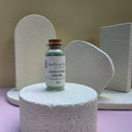 A small glass vial of Healing Hearts & Crafts Paint Pigment Powder in Earthy Sage sits on a textured white cylindrical pedestal. The background features abstract, textured white geometric shapes against a beige wall, with the vial placed on a lavender-pink surface.