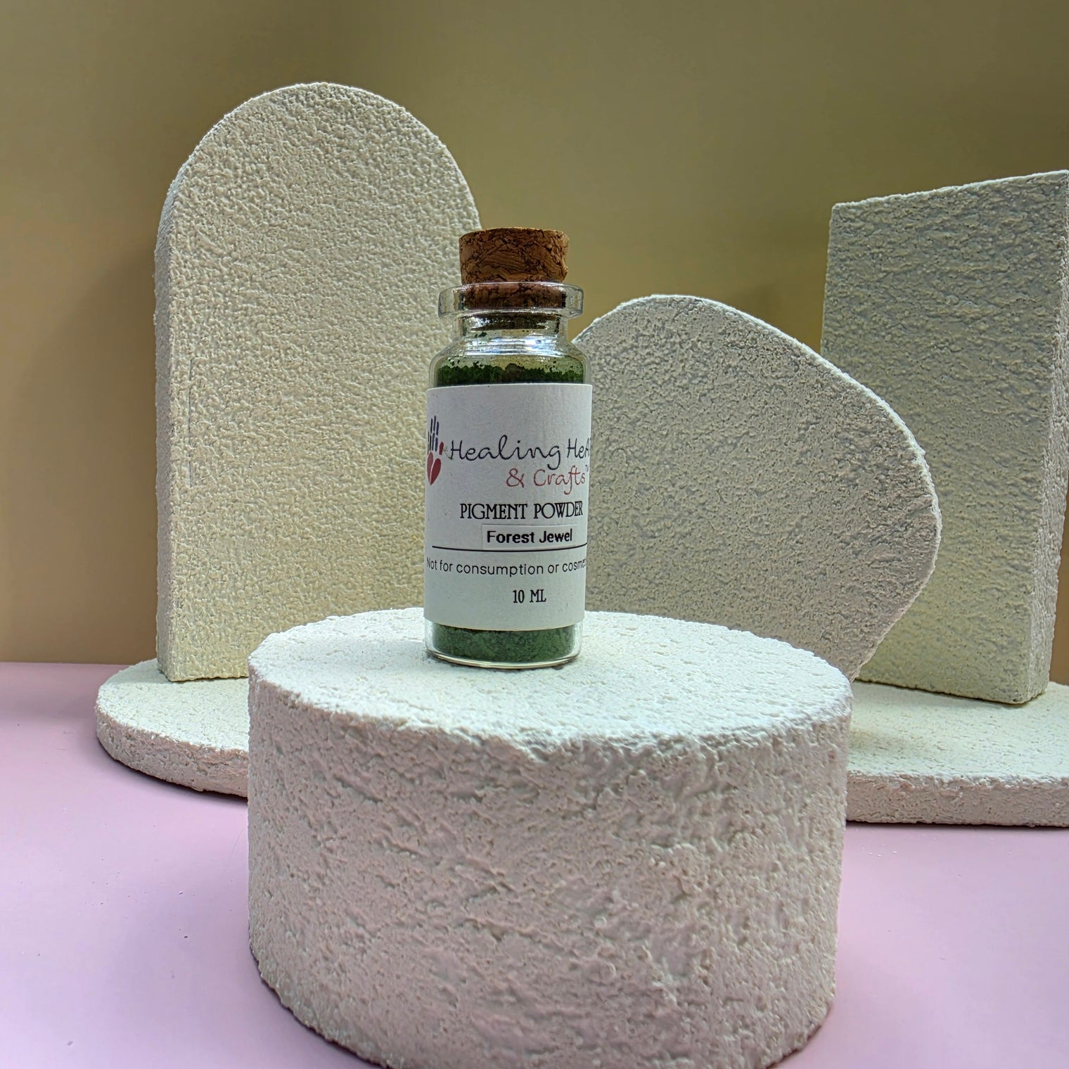 A small glass bottle labeled Healing Hearts & Crafts Paint Pigment Powder in Forest Jewel Green, set on a textured white cylindrical pedestal with sculptural props in the background.