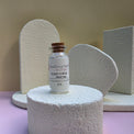 A small glass vial labeled Healing Hearts & Crafts Pigment Powder in Glacier Dust with a cork stopper, displayed on a textured white pedestal against a beige and lavender studio background.