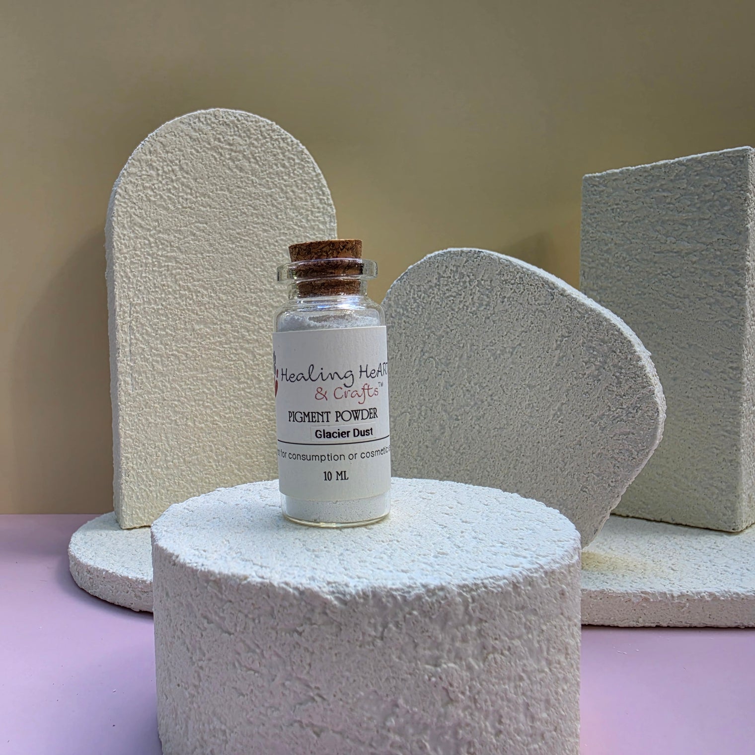 A small glass vial labeled Healing Hearts & Crafts Pigment Powder in Glacier Dust with a cork stopper, displayed on a textured white pedestal against a beige and lavender studio background.