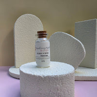 A small glass vial labeled Healing Hearts & Crafts Pigment Powder in Glacier Dust with a cork stopper, displayed on a textured white pedestal against a beige and lavender studio background.