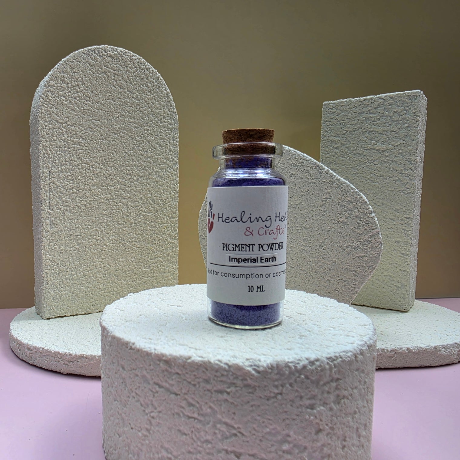 A small glass vial of paint pigment powder by Healing Hearts & Crafts in Imperial Earth sits on a textured white cylindrical pedestal. The vial contains deep purple pigment powder and has a cork stopper.