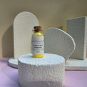 A small glass vial of Healing Hearts & Crafts  Paint Pigment Powder in Luminous Field sits on a textured white cylindrical pedestal. The vial contains a bright yellow powder and has a cork stopper.