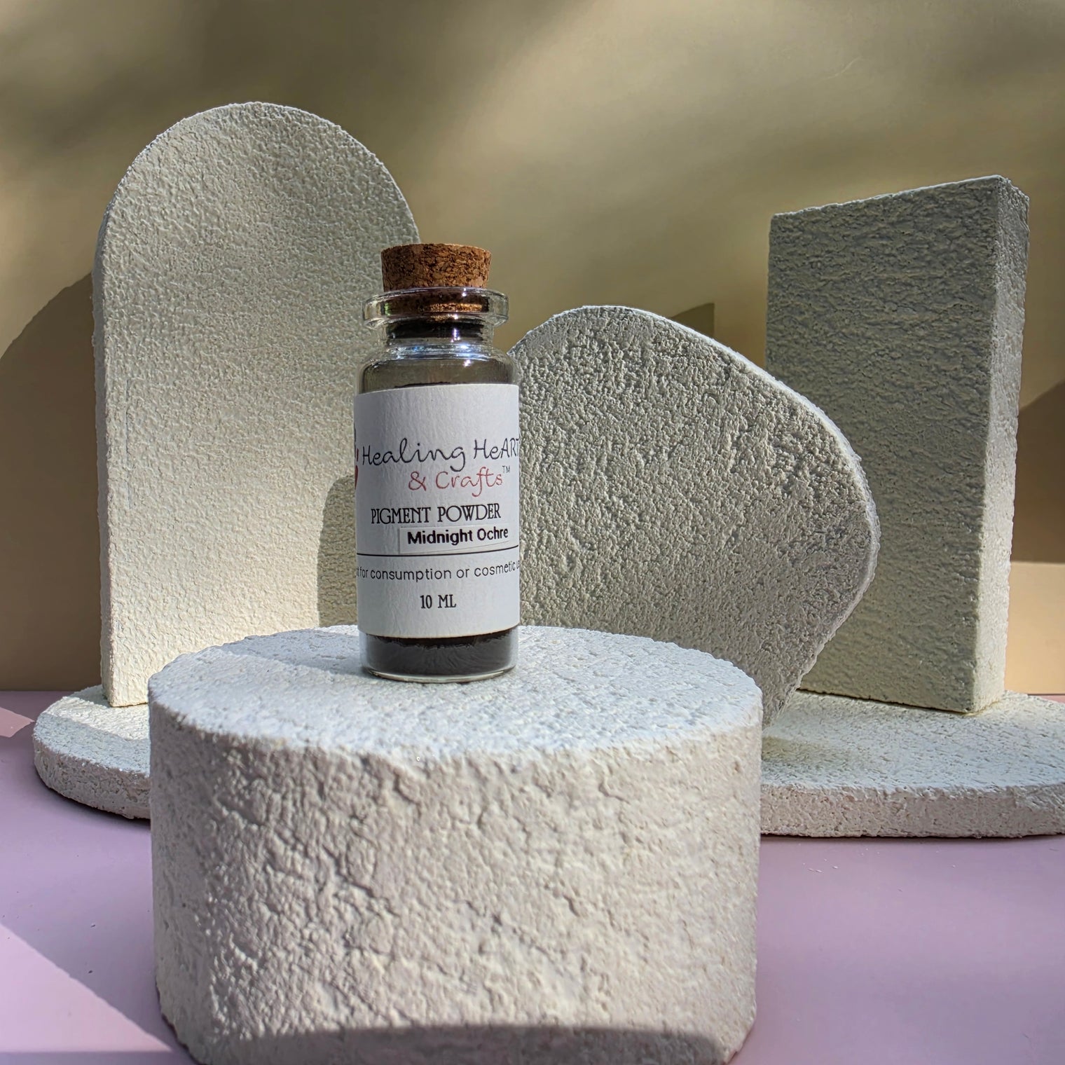 A small glass bottle of Healing Hearts & Crafts Midnight Ochre Paint Pigment powder sits on a textured white pedestal. The bottle has a cork stopper and a white label with black and pink text.