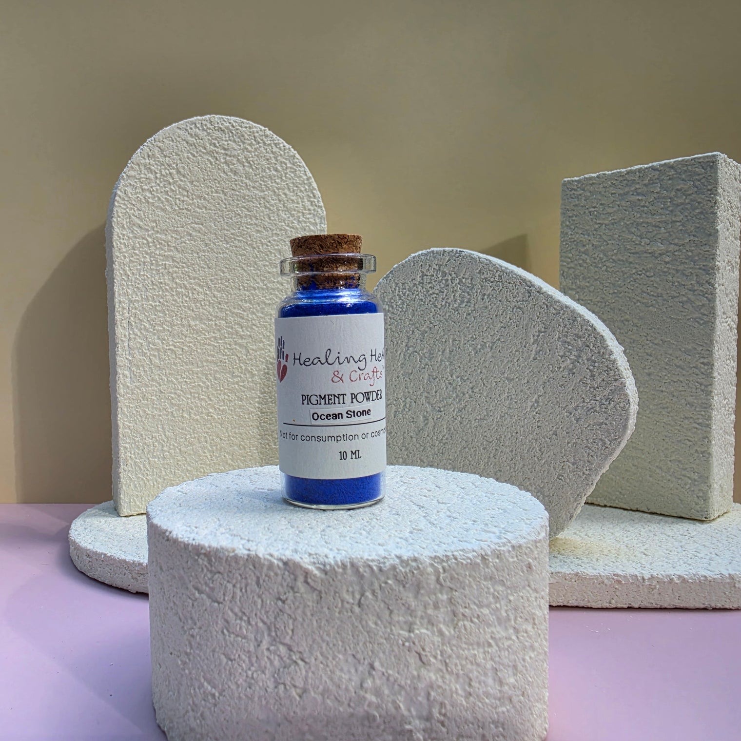 A small glass vial of Healing Hearts & Crafts Paint Pigment Powder in Ocean Stone sits on a textured white cylindrical pedestal. The vial has a cork stopper and a white label with pink and blue text.