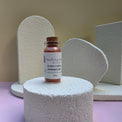A small glass vial of Healing Hearts & Crafts Paint Pigment Powder in Renaissance Earth sits on a textured white cylindrical pedestal. The vial has a cork stopper and a white label with pink and blue text.