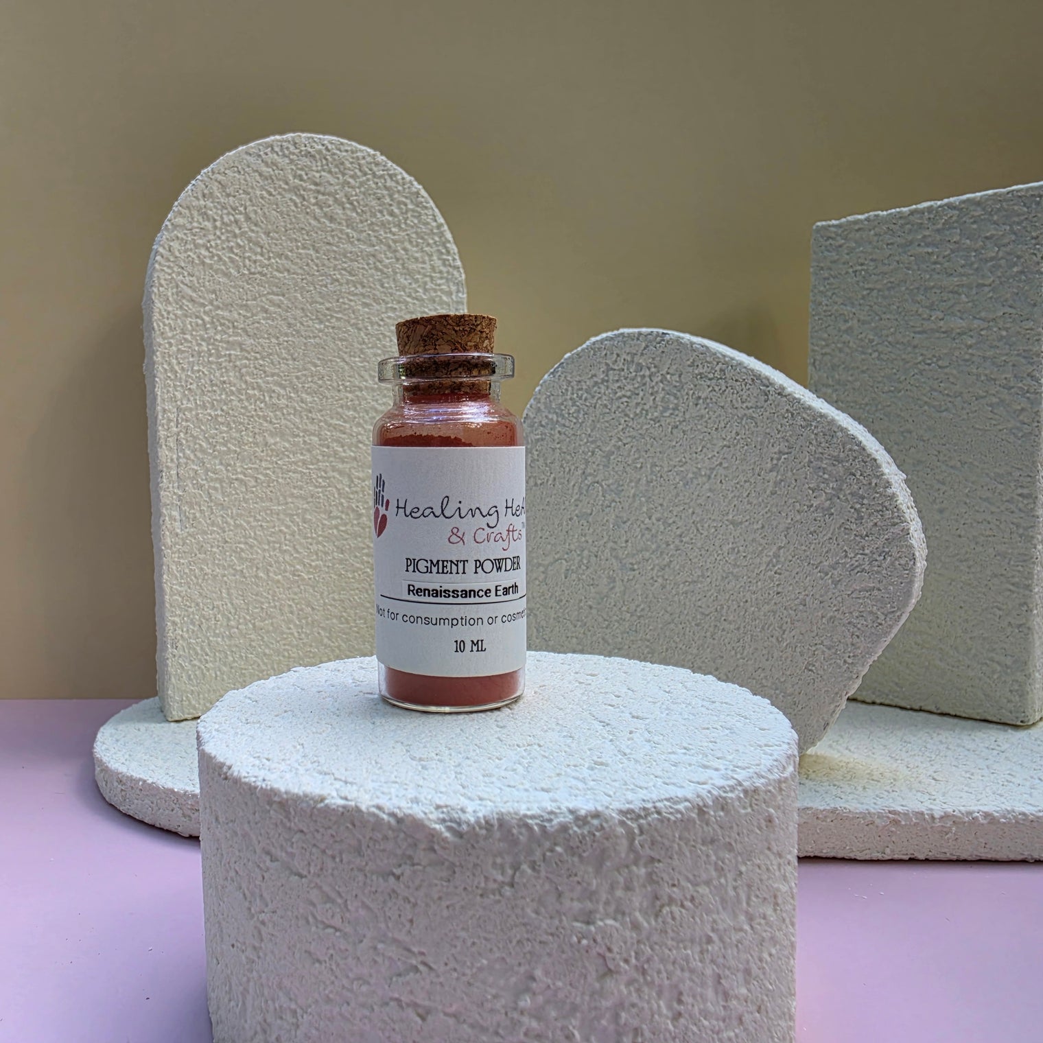 A small glass vial of Healing Hearts & Crafts Paint Pigment Powder in Renaissance Earth sits on a textured white cylindrical pedestal. The vial has a cork stopper and a white label with pink and blue text.