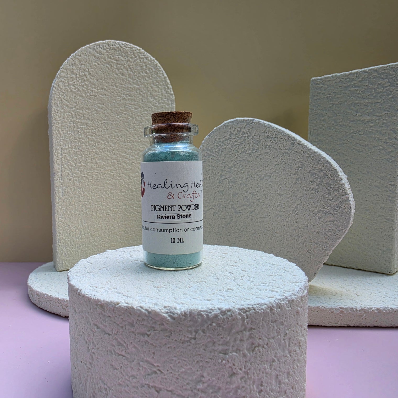 A small glass vial of Healing Hearts & Crafts Rivera Stone Paint pigment powder sits on a textured white cylindrical pedestal. The vial contains a shimmering light blue-green powder and is sealed with a cork stopper, featuring a white label with black text.