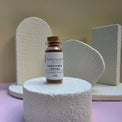 A small glass vial of Healing Hearts & Crafts Rust Ember paint pigment powder sits on a textured white pedestal, with decorative white foam shapes in the background against a beige wall and pink surface.