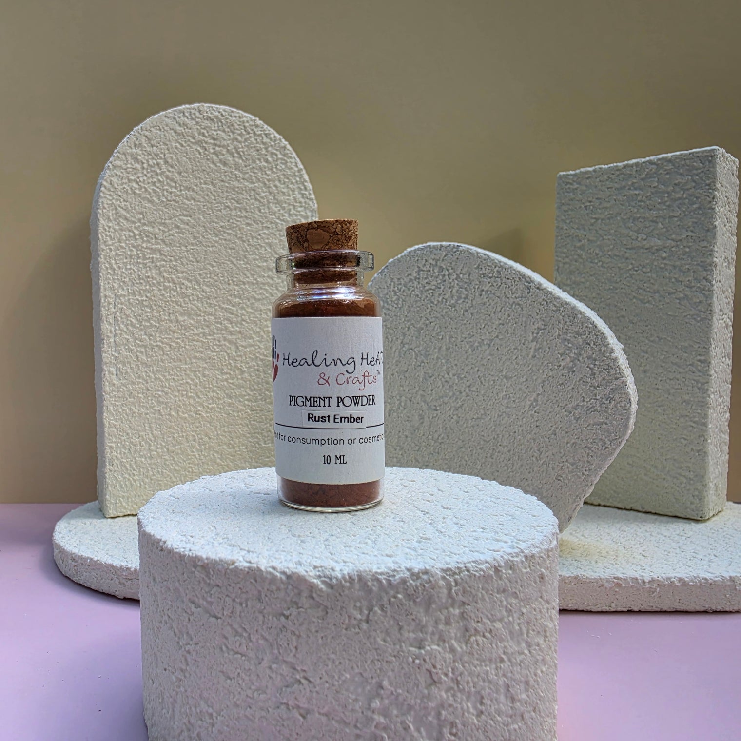 A small glass vial of Healing Hearts & Crafts Rust Ember paint pigment powder sits on a textured white pedestal, with decorative white foam shapes in the background against a beige wall and pink surface.