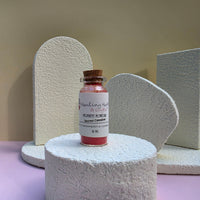 A small glass vial of Healing Hearts & Crafts pigment powder sits on a textured white cylindrical pedestal. The vial contains a vibrant pink pigment powder called Sacred Cinnabar and is sealed with a brown cork stopper featuring a white label.