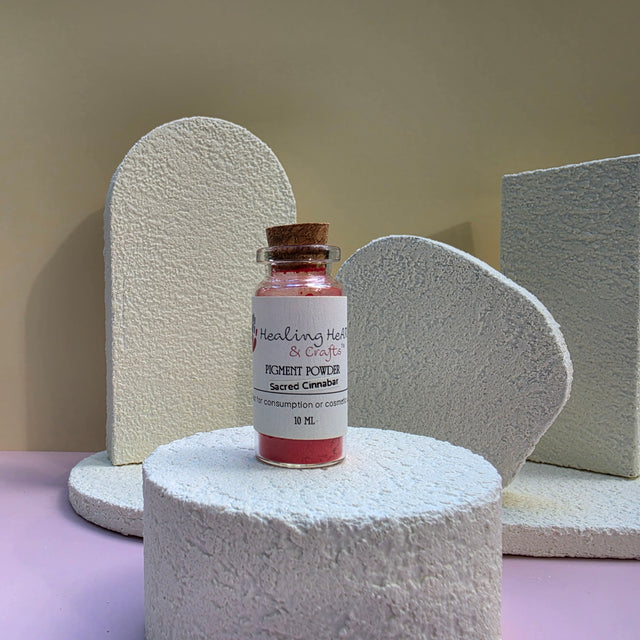 A small glass vial of Healing Hearts & Crafts pigment powder sits on a textured white cylindrical pedestal. The vial contains a vibrant pink pigment powder called Sacred Cinnabar and is sealed with a brown cork stopper featuring a white label.
