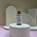 A small glass bottle of Healing Hearts & Crafts Pigment Powder in Smoked Walnut stands on a round textured pedestal, with matching textured display blocks behind it against a beige background.