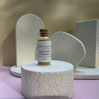 A small glass vial of Healing Hearts & Crafts Pigment Powder in Tuscan Earth sits on a textured white pedestal, surrounded by matching white sculptural props against a beige and pink background.
