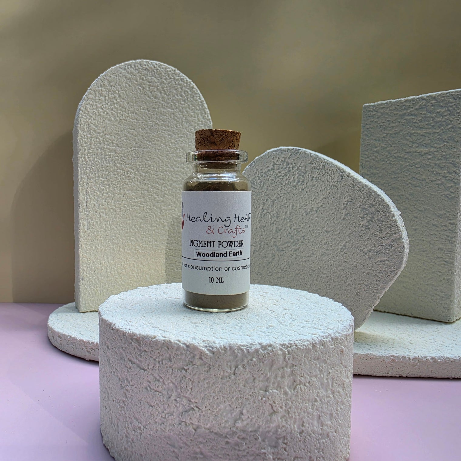 A small glass bottle of Healing Hearts & Crafts Paint Pigment Powder in Woodland Earth is displayed on a textured white cylindrical pedestal, with matching textured white geometric props in the background.