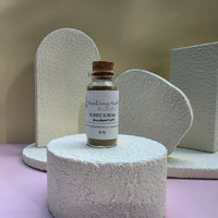 A small glass bottle of Healing Hearts & Crafts Paint Pigment Powder in Woodland Earth is displayed on a textured white cylindrical pedestal, with matching textured white geometric props in the background.